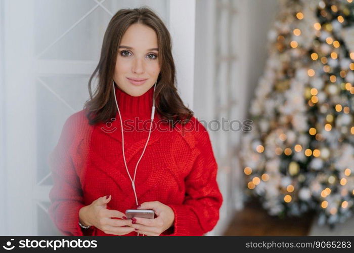 Horizontal shot of young Caucasian woman enjoys music from playlist, listens favourite song with earphones, dressed in knitted red sweater, stands against decorated Christmas tree background