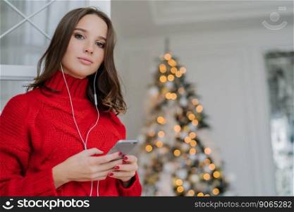 Horizontal shot of young European girl has make up, healthy skin, dark hair, dressed in oversized sweater, listens audio book with earphones and cell phone, poses near decorated Christmas tree
