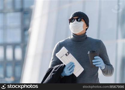 Horizontal shot of young man in medical mask prevents infectious pneumonia, coronavirus pandemic, virus epidemic, drinks coffee to go, going to work, poses outside, has thoughtful expression