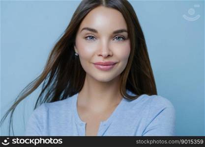 Horizontal shot of young woman with straight dark hair, fresh look and makeup, cares about her skin satisfied with skin care product leaves positive comment poses at studio indoor. Beauty, cosmetology