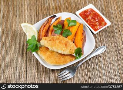 Horizontal top view photo of fried golden breaded coated fish, yams and salsa sauce in white plates with bamboo place mat underneath
