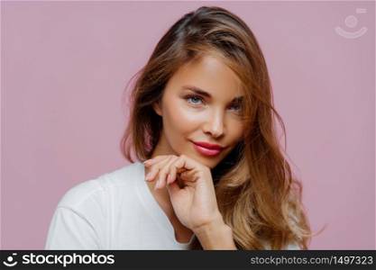 Horizontal view of attractive young woman with long hair, manicure and makeup, looks confidently at camera, wears white sweater, poses against purple background, enjoys spare time. Beauty concept