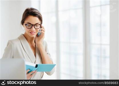 Horizontal view of business lady discusses working schedule with partner via modern smartphone, makes solution for project, focused in notepad, wears spectacles and formal suit, stands indoor