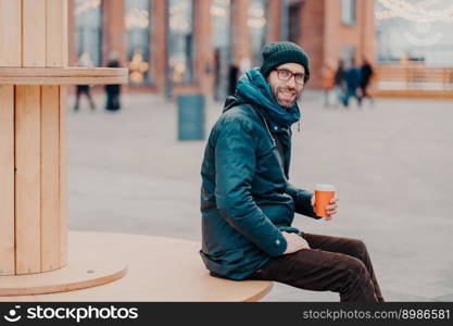 Horizontal view of cheerful European man with thick bristle, wears hat and jacket, holds disposable cup of coffee, breathes fresh air, poses at street. Male model enjoys fresh beverage outside