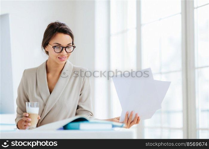 Horizontal view of confident brunette female worker in formal wear and optical spectacles looks through documentation, drink fresh beverage, focused down, poses indoor, has pleased expression