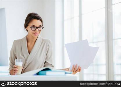 Horizontal view of confident brunette female worker in formal wear and optical spectacles looks through documentation, drink fresh beverage, focused down, poses indoor, has pleased expression