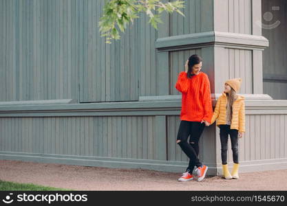 Horizontal view of fashionable European female in red loose sweater, ripped jeans, holds hands together with her younger sister, pose outside together, have walk in open air, have friendly relations