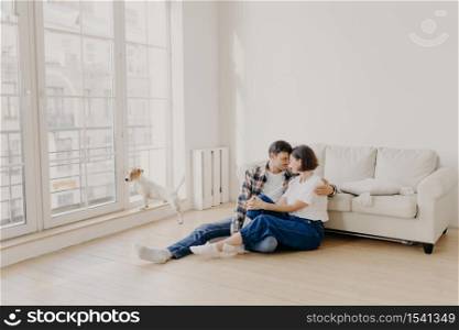 Horizontal view of happy affectionate family couple dressed in casual wear, embrace and express love to each other, pose on floor near sofa in modern apartment, their pet looks through big window