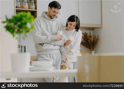 Horizontal view of happy young woman and man play with dog, drink takeaway coffee, stand against kitchen interior, unpack boxes with personal stuff, wear casual clothes, bought new apartment