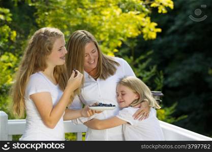 Horizontal view of mother and older sister looking at youngest daughter while eating fresh blue berries outdoors on patio with woods in background
