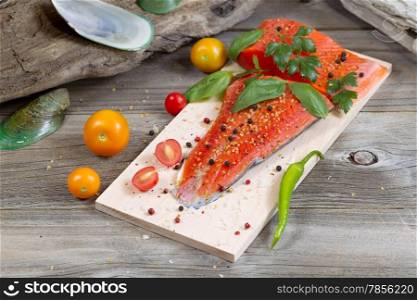 Horizontal view of raw red salmon, skin side down, on maple wood grilling plank with seasoning and other herbs surrounded by drift wood and shells