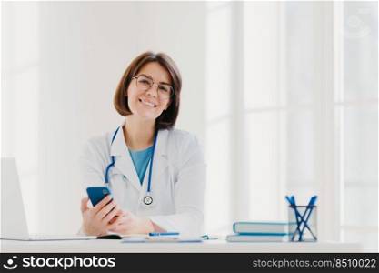 Horizontal view of smiling professional doctor works in clinic, poses at modern hospital office with electronic gadgets, sends text messages on cellphone being at work. Health care, technology concept