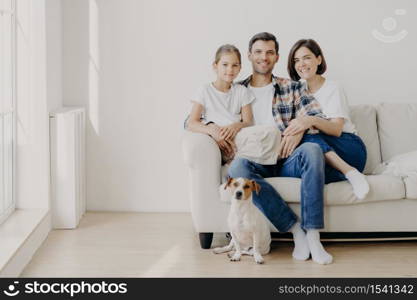 Horizontal view of three friendly family members and small pedigree dog pose at camera for making memorable photography. Dauther spends free time with mother and father, have good day at home