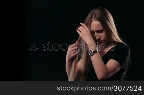 Horrified young beautiful woman with long blonde hair found her first grey hair over black background. Unhappy female looking with open mouth at first grey hair almost ready to cry. Human facial expression, emotion. Beauty hairstyle.