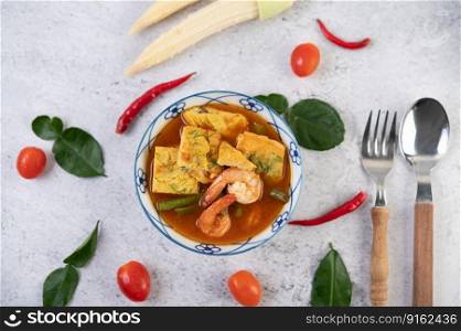 Hot and sour soup with cha-om, egg, and shrimp in a white bowl, with chili and kaffir lime leaves on white background.