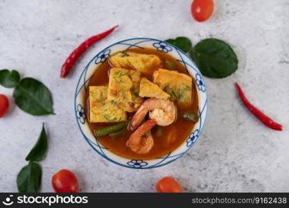 Hot and sour soup with cha-om, egg, and shrimp in a white bowl, with chili and kaffir lime leaves on white background.