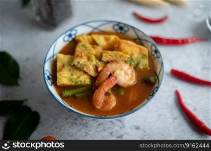 Hot and sour soup with cha-om, egg, and shrimp in a white bowl, with chili and kaffir lime leaves on white background.