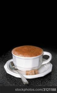Hot cappuccino with spoon and sugar cubes on a dark wood table.