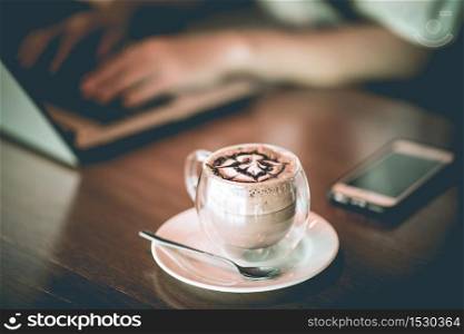 Hot Chocolate on glass in coffee shop cafe with woman working with laptop in background,Selective Focus
