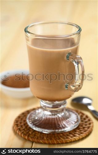 Hot chocolate with cocoa powder and spoon in the back (Selective Focus, Focus on the front rim of the glass)