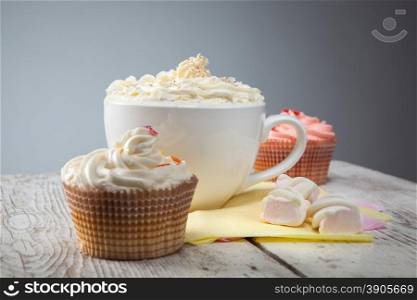 hot chocolate with marshmallows, cream and cupcakes on wooden background