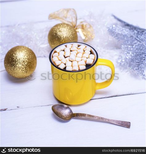 hot chocolate with marshmallows in a yellow cup and Christmas toys on a white wooden background