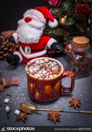 hot cocoa with marshmallow in a brown ceramic mug on a black background, behind a textile Santa Claus and Christmas tree