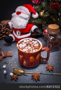 hot cocoa with marshmallow in a brown ceramic mug on a black background, behind a textile Santa Claus and Christmas tree