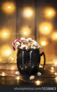 Hot cocoa with marshmallows and whipped cream in a black mug and string lights, on wooden background. Christmas lights and hot drink. Coziness concept