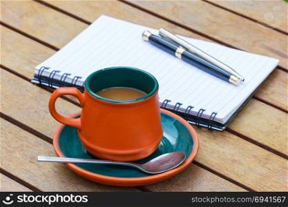 Hot coffee and notebook and pens on the wooden table. Focus on a cup.
