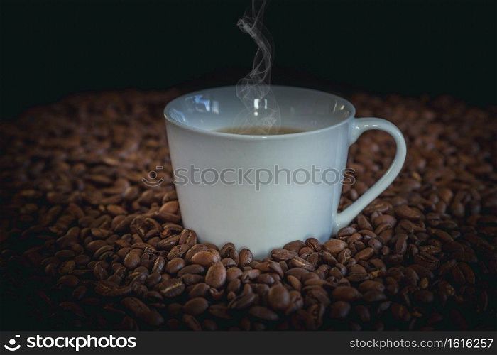Hot coffee in a white coffee cup and many coffee beans placed around and small wooden bucket of coffee on a wooden table in a warm, light atmosphere, on dark background, with copy space.