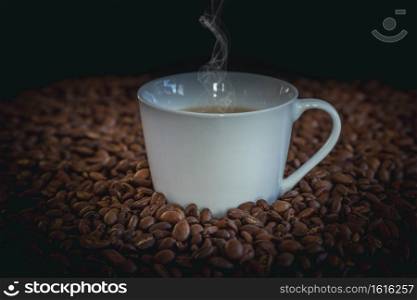 Hot coffee in a white coffee cup and many coffee beans placed around and small wooden bucket of coffee on a wooden table in a warm, light atmosphere, on dark background, with copy space.