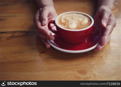 Hot coffee latte cappuccino in red cup and saucer with beautiful latte art milk foam on barista&rsquo;s hands holding serving on wood table background.