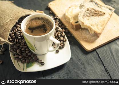 Hot cup of coffee on its plate, surrounded by coffee beans coming out from a small burlap sack and slices of pound cake in background