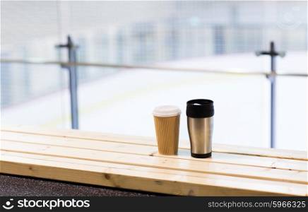 hot drink, winter and leisure concept - close up of coffee cup and thermos on wooden bench at skating rink arena