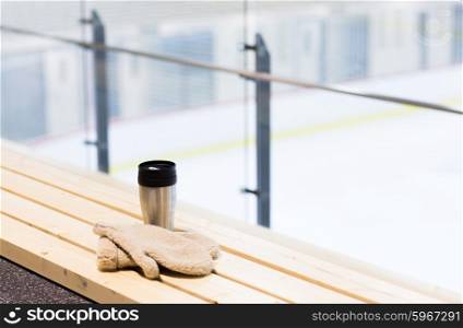hot drink, winter and leisure concept - close up of thermos cup and sheepskin mittens on bench at ice rink arena