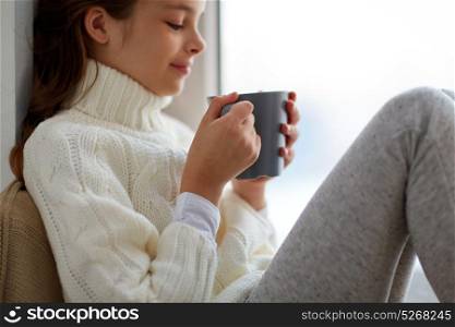 hot drinks and people concept - beautiful girl in winter sweater with tea mug sitting at home window. girl with tea mug sitting at home window