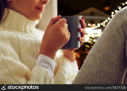 hot drinks, holidays and people concept - close up of beautiful girl in winter sweater with tea mug sitting at window over christmas lights background. close up of girl with tea mug sitting at window