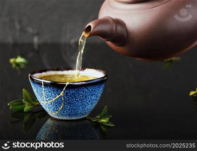 Hot green tea is poured from a ceramic teapot into a bowl. Selective focus on the blue cup. Steam will rise above the mug Reclaimed ceramic blue cup, second life of things, recycling or kintsugi