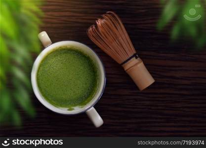 Hot Matcha Green Tea Latte Cup on Wooden Table with Chasen or Bamboo Whisk. Japanese Traditional Drink. Blurred Green Leaf as foreground. Top View