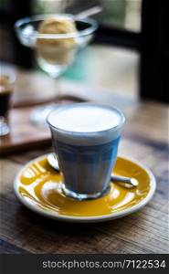 Hot milk with butterfly pea mixed and milk froth on the top of glass, decorate with yellow disk on wooden table in cafe shop mood and tone, selective focus.