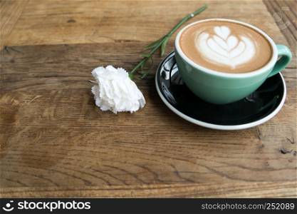 Hot mocha coffee or capuchino in the green cup with heart pattern and white carnation on the wooden table