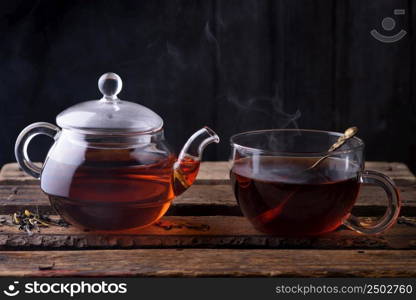 Hot tea in glass teapot and cup with spoon and steam on wooden table dark still life