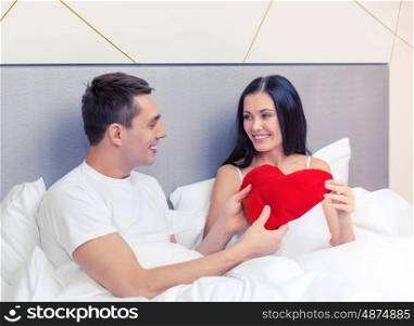hotel, travel, relationships, holidays and happiness concept - smiling couple in bed with red heart-shaped pillow