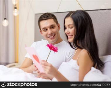 hotel, travel, relationships, holidays and happiness concept - smiling couple in bed with postcard and pink flower