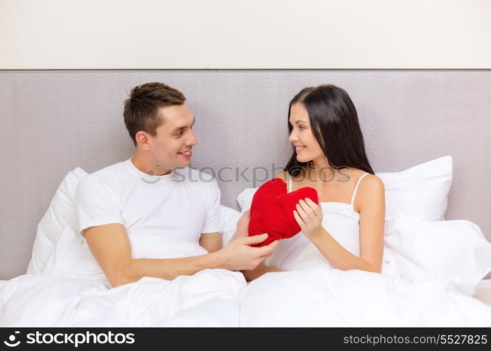 hotel, travel, relationships, holidays and happiness concept - smiling couple in bed with red heart-shaped pillow