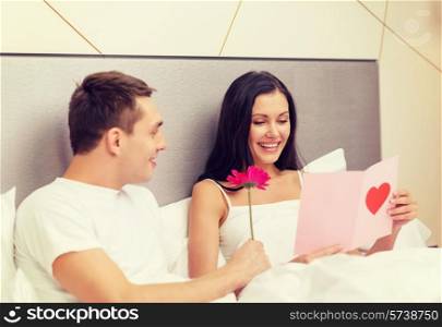 hotel, travel, relationships, holidays and happiness concept - smiling couple in bed with postcard and pink flower