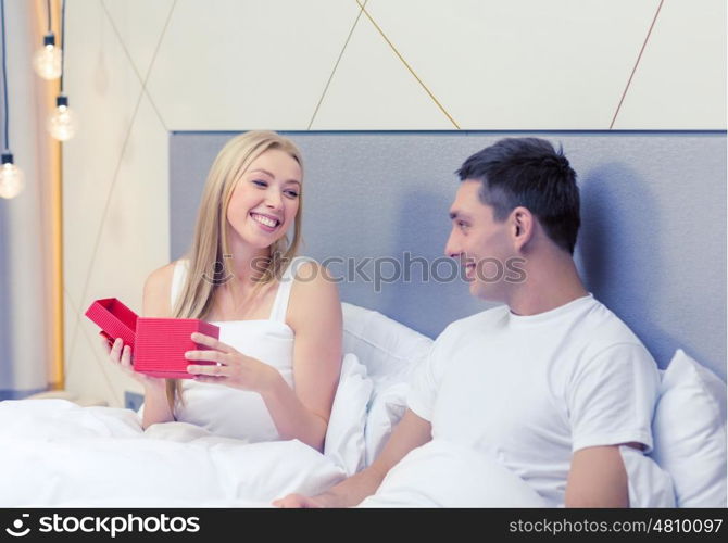 hotel, travel, relationships, holidays and happiness concept - smiling couple in bed with red gift box