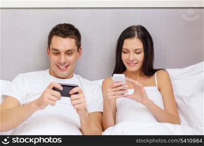 hotel, travel, relationships, technology, intermet and happiness concept - smiling couple in bed with smartphones