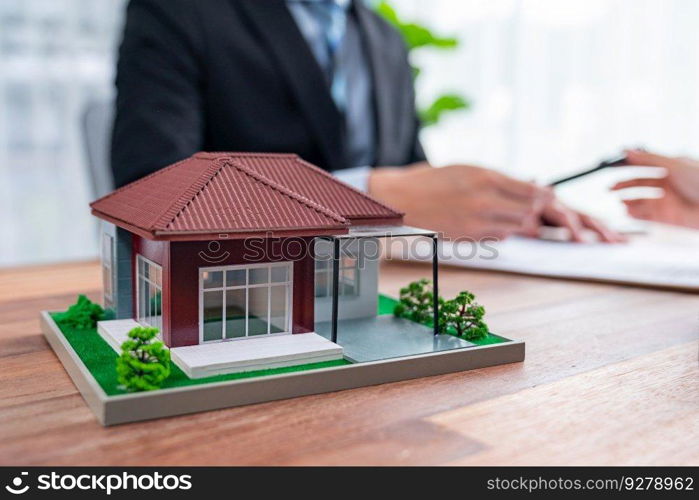 House model sample on wooden table with blurred background of real estate agent and buyer or client discussing terms and condition on house loan contract. Housing business meeting. Jubilant. House model sample on wooden table with blurred background. Jubilant